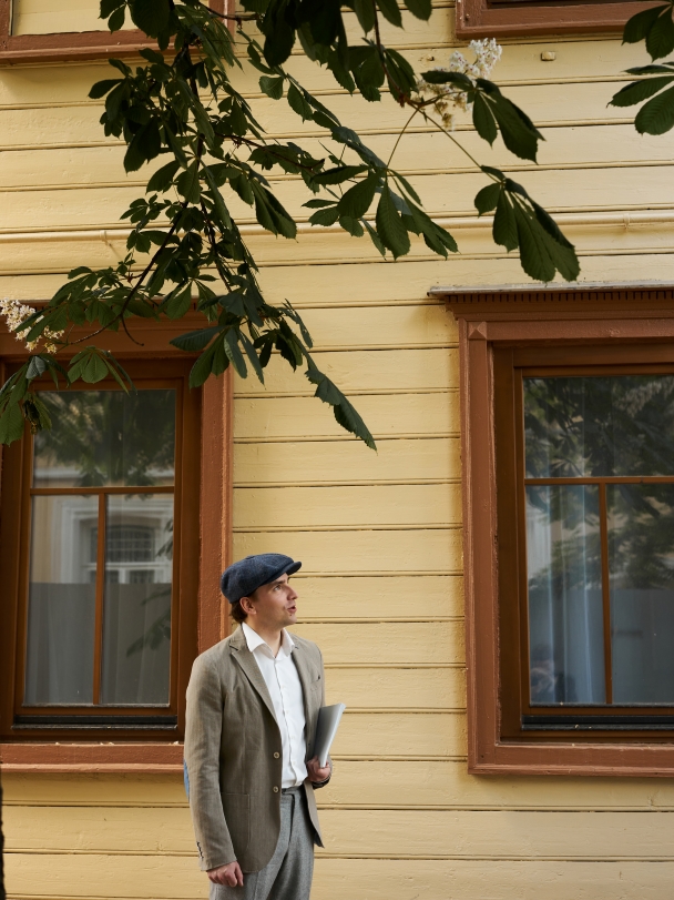 Степан в кепке и льняном пиджаке, с папкой под мышкой; стоит подле окон дома