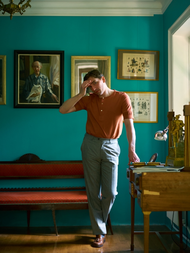 Степан в хенли и серых брюках; стоит, опершись на столик для бумаг, рядом с алым диваном